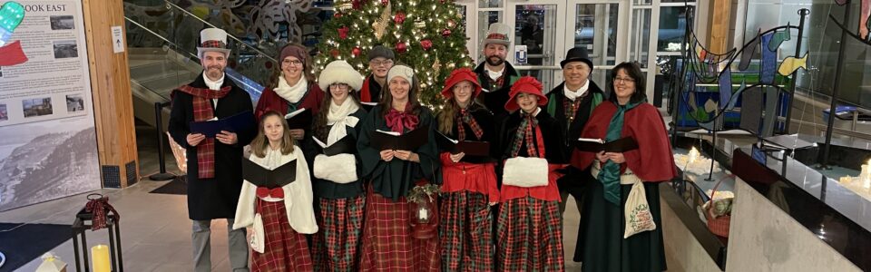 11 adults and kids dressed up in vintage carolling outfits standing in front of a Christmas tree