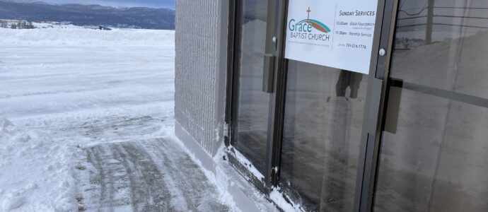 A snowy scene outside a building with a gray concrete exterior and large glass doors. A sign on the door reads "Grace Baptist Church" with service times listed: Bible Foundation at 10:00 a.m. and Worship Service at 11:30 a.m., along with a phone number. Snow covers the ground, sidewalk, and the base of the doors, with a clear view of snow-covered mountains and a frozen landscape in the background under a gray sky.