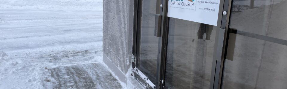 A snowy scene outside a building with a gray concrete exterior and large glass doors. A sign on the door reads "Grace Baptist Church" with service times listed: Bible Foundation at 10:00 a.m. and Worship Service at 11:30 a.m., along with a phone number. Snow covers the ground, sidewalk, and the base of the doors, with a clear view of snow-covered mountains and a frozen landscape in the background under a gray sky.