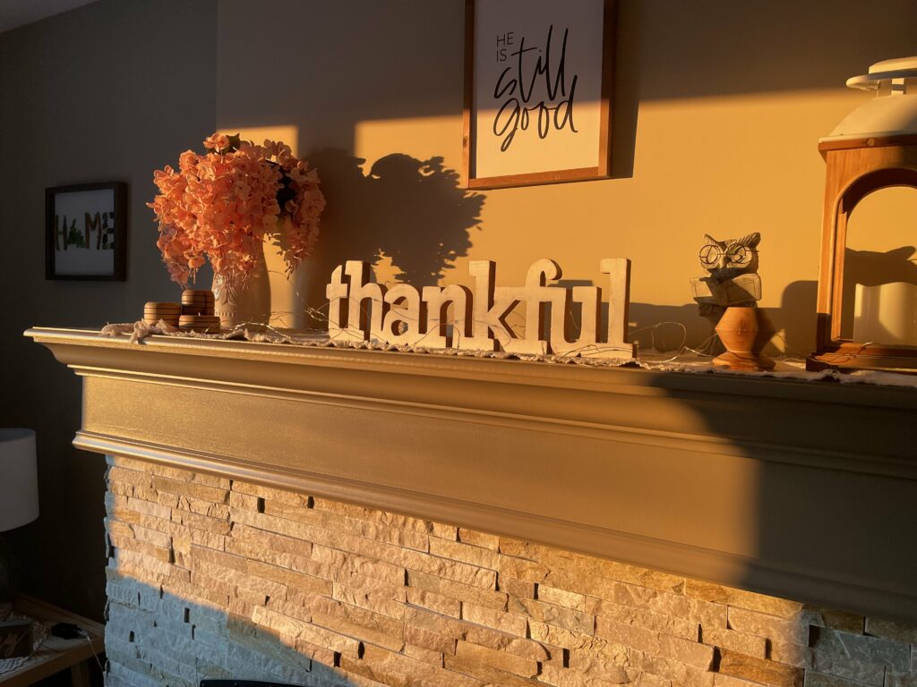 A cozy fireplace mantel adorned with a warm, autumnal decor. The mantel features a large wooden sign spelling out "thankful" in the center. To the left, there is a vase filled with vibrant orange flowers, accompanied by a small stack of round wooden coasters. Above the mantel, a framed sign reads "HE still is good" in elegant handwriting. To the right, a small owl figurine and a decorative lantern with a candle inside are placed. The scene is bathed in soft, golden sunlight, casting gentle shadows and highlighting the stonework of the fireplace below.