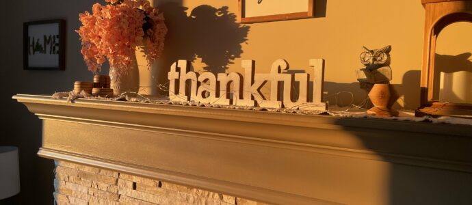 A cozy fireplace mantel adorned with a warm, autumnal decor. The mantel features a large wooden sign spelling out "thankful" in the center. To the left, there is a vase filled with vibrant orange flowers, accompanied by a small stack of round wooden coasters. Above the mantel, a framed sign reads "HE still is good" in elegant handwriting. To the right, a small owl figurine and a decorative lantern with a candle inside are placed. The scene is bathed in soft, golden sunlight, casting gentle shadows and highlighting the stonework of the fireplace below.