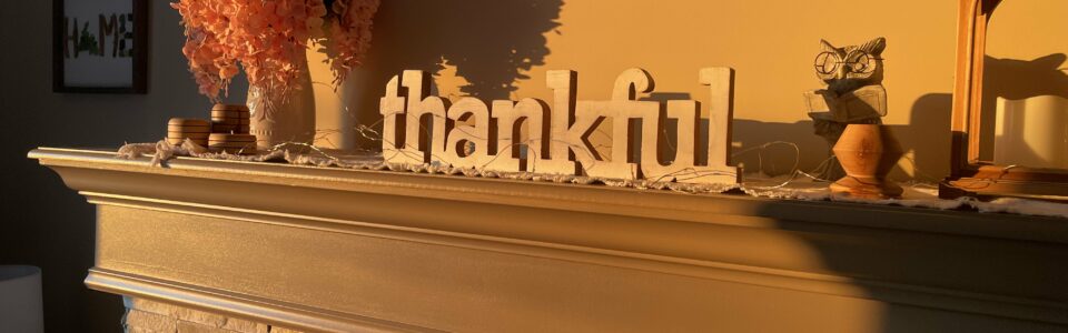 A cozy fireplace mantel adorned with a warm, autumnal decor. The mantel features a large wooden sign spelling out "thankful" in the center. To the left, there is a vase filled with vibrant orange flowers, accompanied by a small stack of round wooden coasters. Above the mantel, a framed sign reads "HE still is good" in elegant handwriting. To the right, a small owl figurine and a decorative lantern with a candle inside are placed. The scene is bathed in soft, golden sunlight, casting gentle shadows and highlighting the stonework of the fireplace below.