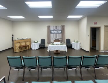 Church sanctuary with a simple pulpit, open Bible on a table, chairs on either side, and a banner reading “How Firm A Foundation” behind beige curtains.