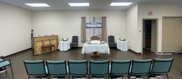 Church sanctuary with a simple pulpit, open Bible on a table, chairs on either side, and a banner reading “How Firm A Foundation” behind beige curtains.