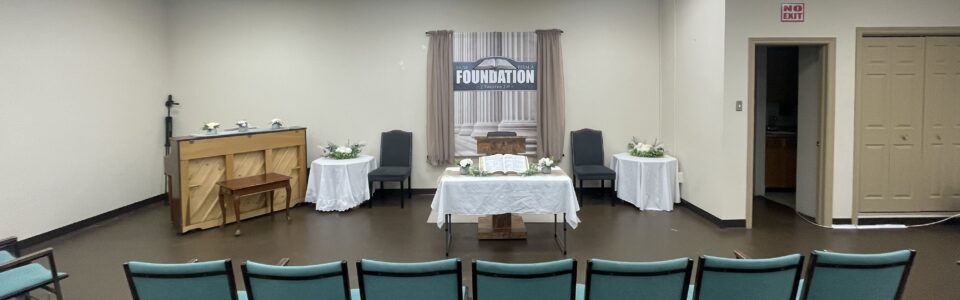 Church sanctuary with a simple pulpit, open Bible on a table, chairs on either side, and a banner reading “How Firm A Foundation” behind beige curtains.